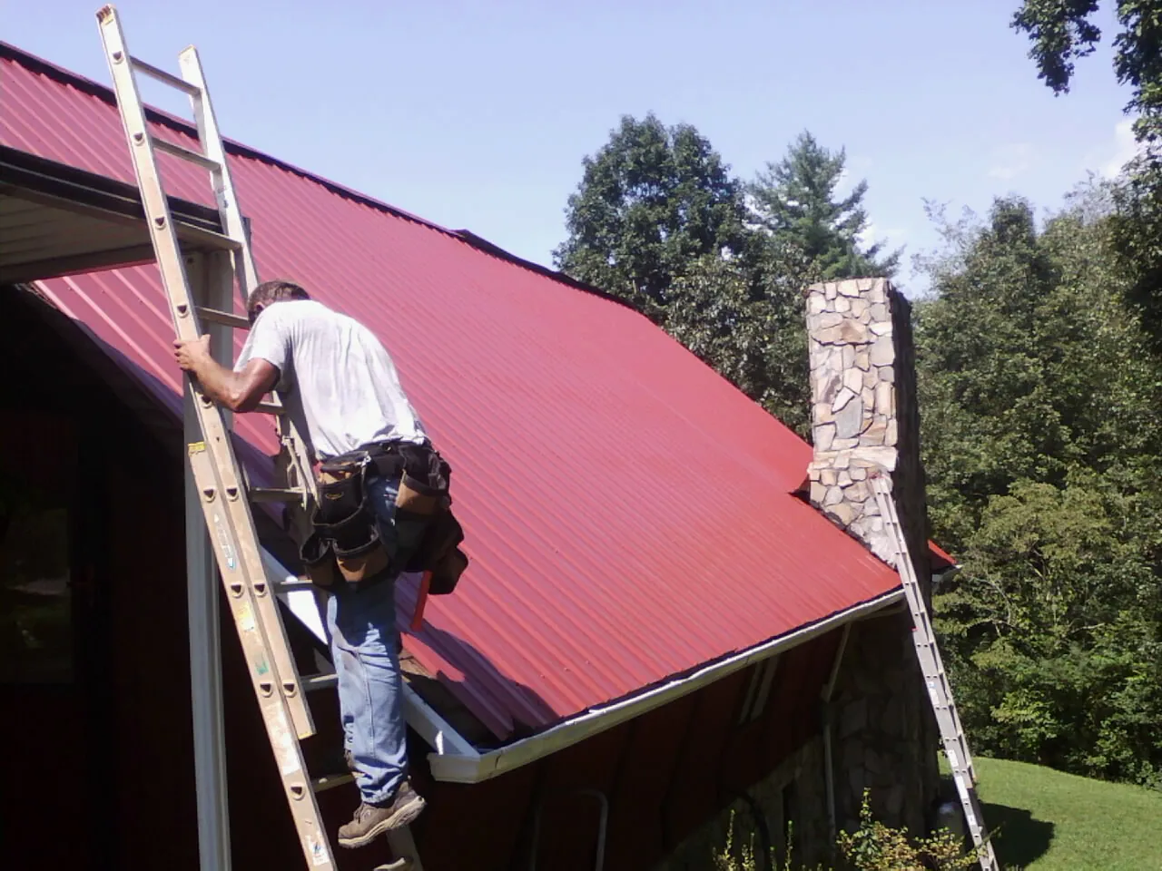 Roofer on a ladder beside a red metal roof and stone chimney — Soffit & Fascia Repair by Roofing Brothers in San Quentin